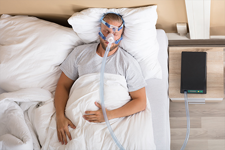 Young Man Lying On Bed With Sleeping Apnea And CPAP Machine